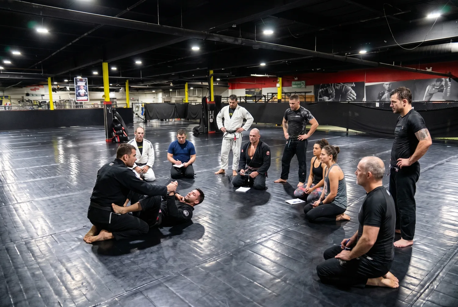 A coach demonstrating a basic technique to a group of attentive adult beginners on the mats